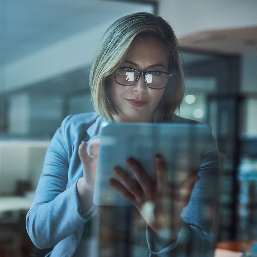 A woman in glasses and a blazer uses a tablet in a dimly lit office, with reflections on her glasses and the tablet screen.
