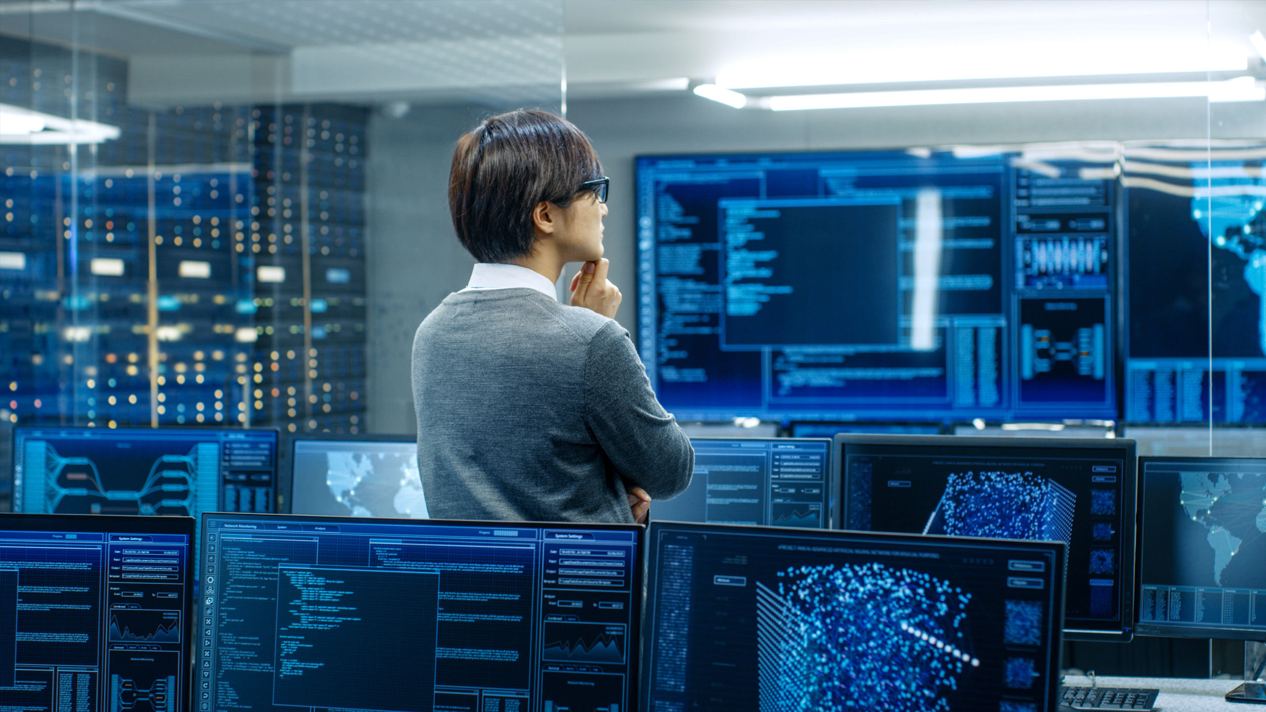 A person in a grey sweater stands in a data center control room, looking at multiple computer monitors displaying code, graphs, and network maps.