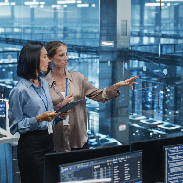 Two women in a data center. One points towards server racks with blue lights, while the other holds a tablet. Computer monitors with code are in the foreground.